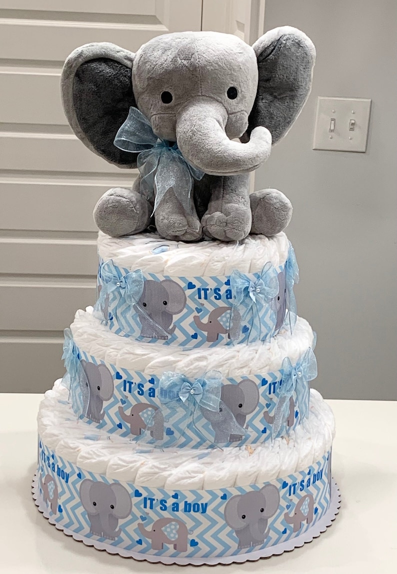 Pode incluir: Um bolo de fraldas de tr&ecirc;s camadas decorado com tecido estampado em chevron azul e branco e aplica&ccedil;&otilde;es de elefante cinza. A camada superior apresenta um elefante de pel&uacute;cia cinza com uma fita azul.