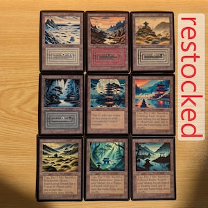 May include: Nine Magic: The Gathering cards with landscape art in a Japanese style. Each card features a different scene, including mountains, forests, and temples. The cards have text at the bottom, and a "restocked" sign is visible.