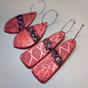 May include: Two pairs of dangle earrings featuring a metallic red finish with embossed floral and geometric patterns. Each earring has a central diagonal band of dark, shimmering, crushed-texture material and a thin silver-toned metal hoop.