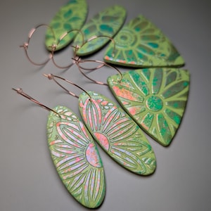 Puede incluir: Una colección de cinco pendientes con formas planas, texturizadas y de color verde claro, con patrones florales en relieve resaltados por acentos metálicos en rosa y plata. Cada pieza está unida a un fino aro de metal de color cobre.