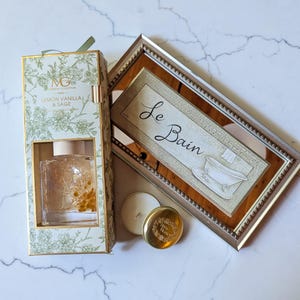 May include: A decorative set featuring a framed mirror with "Le Bain" text, a glass diffuser with dried flowers, and two candles. The diffuser box is labeled "Lemon Vanilla & Sage". The mirror has a brown frame.