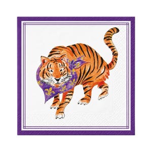 May include: A square paper napkin with a white center and a purple border. The center features a watercolor illustration of an orange tiger with black stripes wearing a purple bandana decorated with yellow fleur-de-lis symbols.