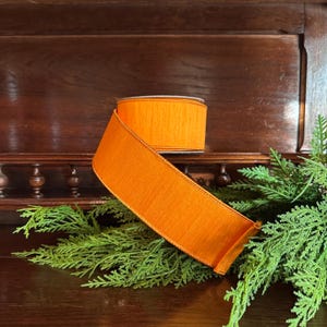 Puede incluir: Un rollo de cinta naranja brillante con una superficie texturizada. La cinta se muestra sobre una superficie de madera oscura con follaje verde. La cinta es un elemento decorativo ancho.