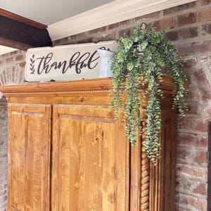May include: A wooden cabinet with a light brown finish, topped with a white sign that reads "thankful" in black script. A hanging plant with green leaves and a black metal lantern are on top of the cabinet. The background is a brick wall.