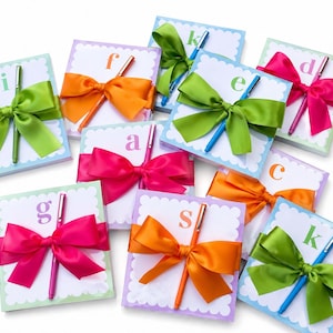 May include: A collection of square notepads with scalloped edges, each adorned with a large satin bow and a pen. The bows are in various colors, including green, pink, and orange. Each notepad has a letter printed on it.