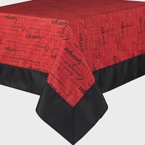 May include: A red tablecloth with the word "Georgia" printed repeatedly in black script. The tablecloth has a black border and is draped over a table. The overall theme is a state-themed home decor item.