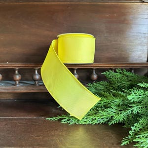 May include: A bright yellow satin ribbon spool, perfect for crafting and gift wrapping. The ribbon is displayed on a wooden surface with green foliage.
