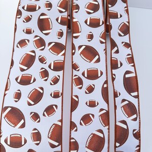 May include: A white ribbon featuring a repeating pattern of brown American footballs with white laces. The ribbon has finished edges in a solid brown color, creating a distinct border along the length of the fabric strips displayed vertically.