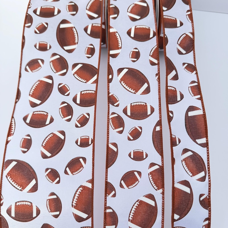May include: A white ribbon featuring a repeating pattern of brown American footballs with white laces. The ribbon has finished edges in a solid brown color, creating a distinct border along the length of the fabric strips displayed vertically.