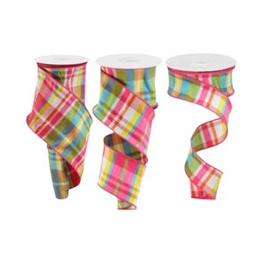 May include: Three spools of colorful plaid ribbon. The ribbon features a pattern of pink, red, yellow, green, and blue squares. The edges of the ribbon are finished with a pink trim. The spools are white.