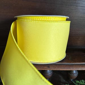 May include: A spool of bright yellow satin ribbon with a white edge. The ribbon is rolled up and ready to be used for crafting or decorating.