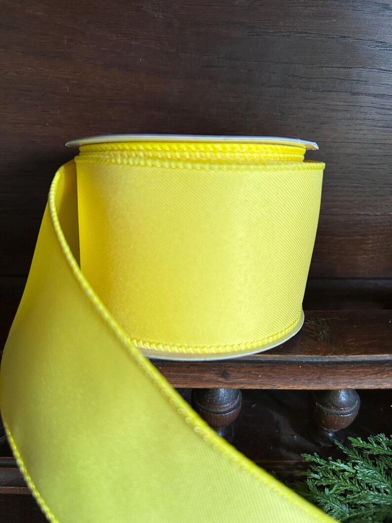May include: A spool of bright yellow satin ribbon with a white edge. The ribbon is rolled up and ready to be used for crafting or decorating.
