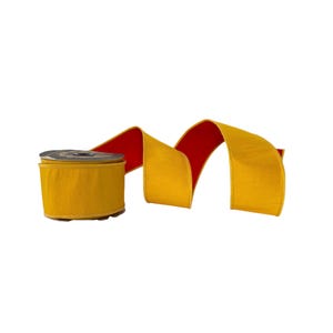 May include: A spool of wide ribbon featuring a vibrant mustard yellow exterior and a contrasting deep red interior. The ribbon is wound around a dark circular core and extends outward in a series of soft, flowing curves, showcasing the dual-colored fabric.