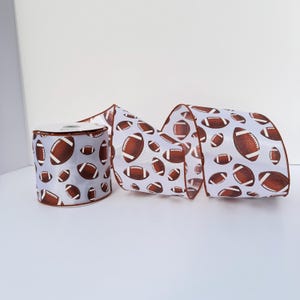 May include: A roll of white craft ribbon featuring a repeating pattern of brown American footballs with white laces. The ribbon has a smooth texture and is displayed as a single spool with a portion of the ribbon unrolled to show the print.