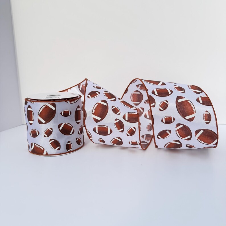 May include: A roll of white craft ribbon featuring a repeating pattern of brown American footballs with white laces. The ribbon has a smooth texture and is displayed as a single spool with a portion of the ribbon unrolled to show the print.