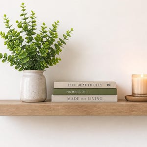 May include: A wooden shelf displays a vase with green foliage, three books with titles "LIVE BEAUTIFULLY", "HOMEBODY", and "MADE FOR LIVING", and a lit candle in a glass holder. The candle sits on a wooden coaster.
