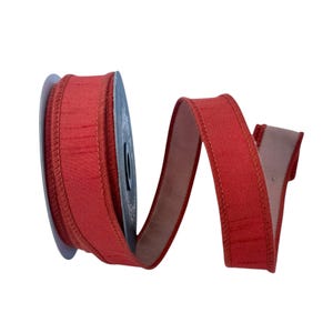 May include: A spool of solid red ribbon with a textured fabric surface and stitched edges. The ribbon is wound around a circular core and extends outward in a loose, flowing S-shaped curve, displaying a consistent deep red color throughout.