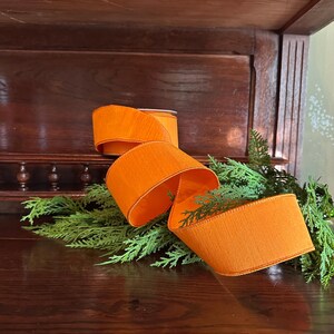Puede incluir: Cinta naranja brillante, desenrollada y rizada, descansa sobre un lecho de follaje verde. La cinta es gruesa y parece estar hecha de una tela texturizada. El fondo es una superficie de madera oscura.
