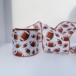 May include: A roll of white ribbon featuring a repeating pattern of brown American footballs with white laces. The ribbon has a finished brown edge along both sides, and the material appears smooth and slightly reflective in the light.