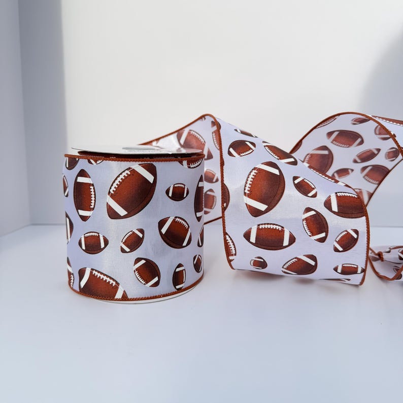 May include: A roll of white ribbon featuring a repeating pattern of brown American footballs with white laces. The ribbon has a finished brown edge along both sides, and the material appears smooth and slightly reflective in the light.