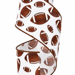 May include: A roll of white wired ribbon featuring a repeating pattern of brown American footballs with white laces. The ribbon has a dark brown stitched edge along both sides, providing a defined border to the fabric material throughout the roll.