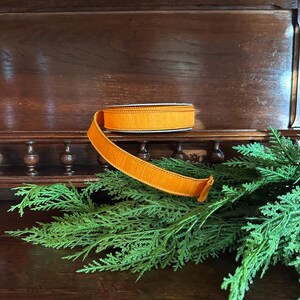 Puede incluir: Un rollo de cinta naranja con un borde texturizado y deshilachado descansa sobre una estanter&iacute;a de madera. La cinta est&aacute; parcialmente desenrollada, revelando su color y textura. El follaje verde est&aacute; en primer plano, con un mueble de madera oscura en el fondo.