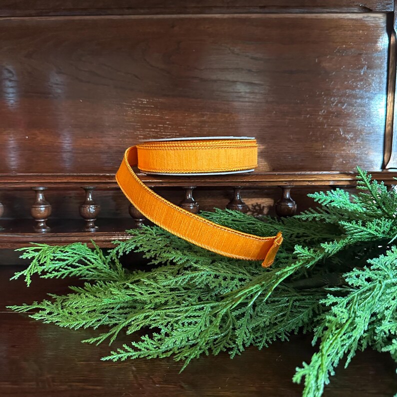 Puede incluir: Un rollo de cinta naranja con un borde texturizado y deshilachado descansa sobre una estanter&iacute;a de madera. La cinta est&aacute; parcialmente desenrollada, revelando su color y textura. El follaje verde est&aacute; en primer plano, con un mueble de madera oscura en el fondo.