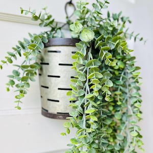 May include: A galvanized metal hanging planter with a distressed finish, filled with cascading faux eucalyptus greenery. The planter has a dark rim and a series of horizontal slots. The leaves are a vibrant green, creating a natural aesthetic.