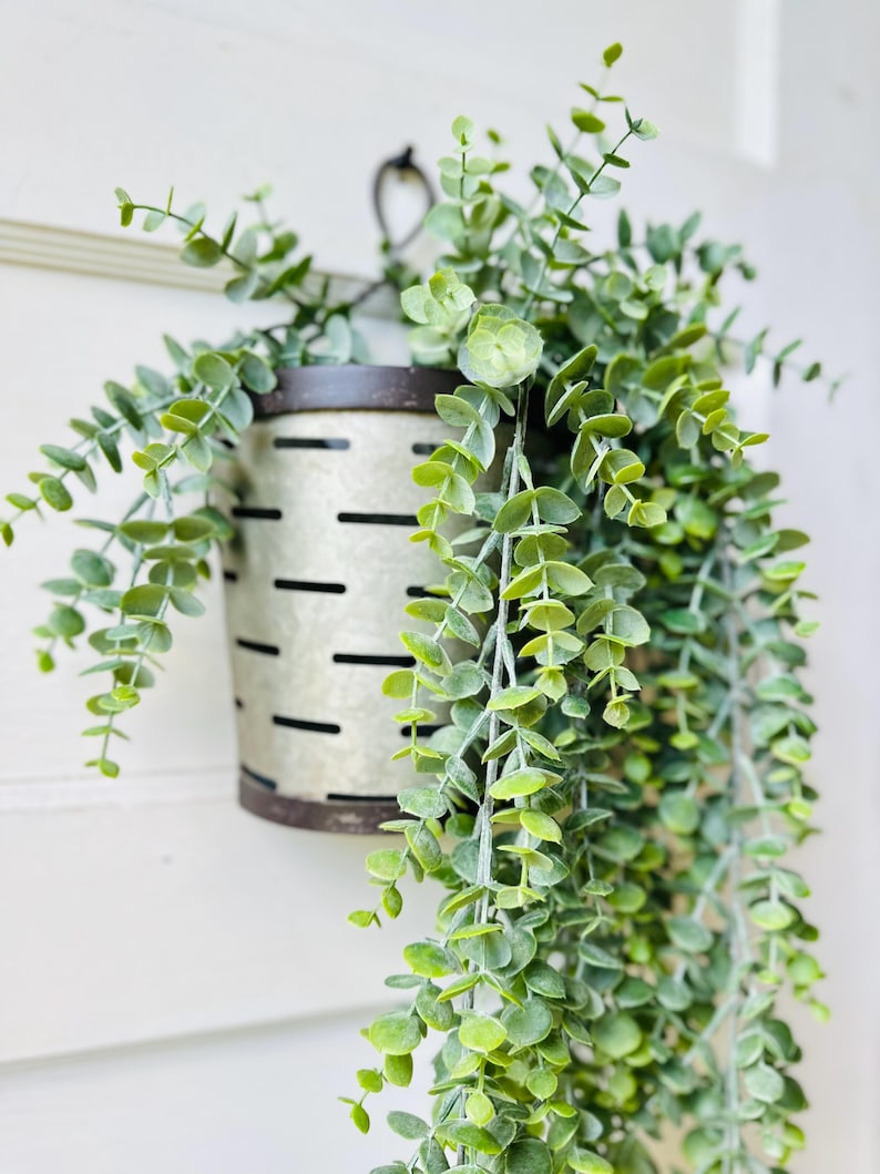 May include: A galvanized metal hanging planter with a distressed finish, filled with cascading faux eucalyptus greenery. The planter has a dark rim and a series of horizontal slots. The leaves are a vibrant green, creating a natural aesthetic.