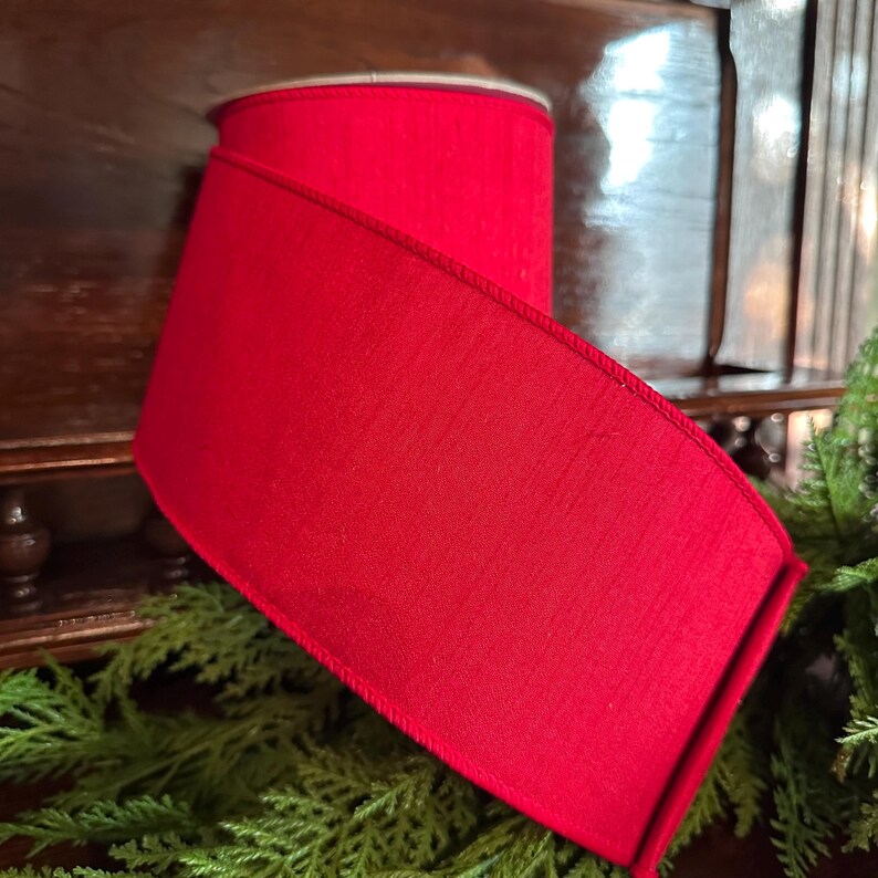 Puede incluir: Un rollo de cinta roja vibrante, perfecto para manualidades o envolver regalos. La cinta tiene una superficie texturizada y un borde acabado. La cinta se muestra sobre una superficie de madera con follaje verde.