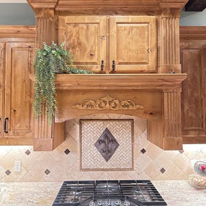 May include: A custom kitchen range hood with a decorative fleur-de-lis tile backsplash. The hood is made of light-colored wood with ornate carvings and a built-in cabinet. A stainless steel gas cooktop is visible below.