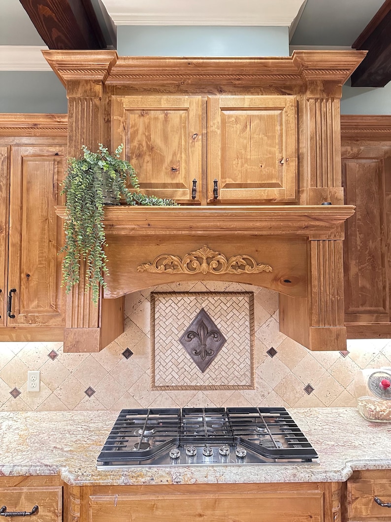 May include: A custom kitchen range hood with a decorative fleur-de-lis tile backsplash. The hood is made of light-colored wood with ornate carvings and a built-in cabinet. A stainless steel gas cooktop is visible below.