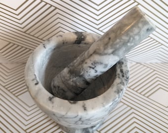 May include: A marble mortar and pestle set. The mortar is a round bowl with a matching pestle. The set is white with gray veining. The pestle is inside the mortar.