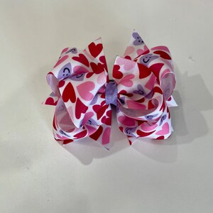 May include: A large, decorative hair bow featuring a pattern of red, pink, and purple hearts on a white background. The bow is tied with a lavender ribbon in the center. The hearts have smiling faces.