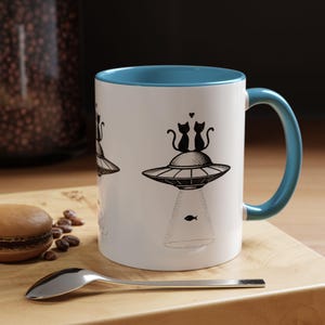 May include: White ceramic mug with a blue rim and handle. The mug features a black and white illustration of two cats riding in a flying saucer. The saucer is emitting a beam of light that ends in a fish.