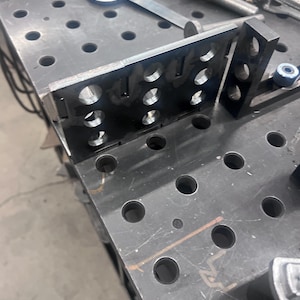 Puede incluir: Primer plano de una mesa de fabricación de metal gris oscuro con numerosos agujeros redondos. Un soporte metálico con varios agujeros circulares se coloca sobre la mesa, junto con otras herramientas y componentes metálicos, lo que sugiere un taller o un entorno industrial.