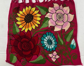 May include: A pink fabric tote bag with embroidered flowers in shades of red, yellow, blue, green, and pink. The bag has a pink rope handle.