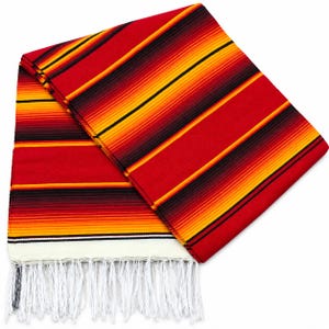 May include: A folded rectangular blanket featuring a vibrant striped pattern in shades of red, orange, yellow, and black. The fabric displays horizontal bands of varying widths and colors, finished with a white fringe along the bottom edge.