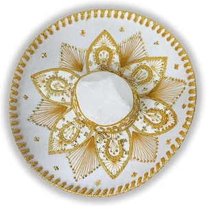 Op de afbeelding: Een witte sombrero met ingewikkelde gouden borduursels en afwerking. De hoed heeft een centrale kroon en een brede, cirkelvormige rand versierd met gedetailleerde patronen en versieringen. Het ontwerp bevat bloemen en geometrische motieven.