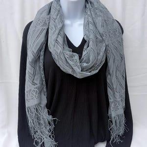 May include: A light gray scarf featuring an intricate openwork lace pattern with a chevron design and long fringe at both ends. The scarf is draped around a white mannequin wearing a black long-sleeve shirt with a textured waffle knit fabric.