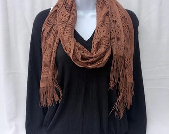 May include: A long, rust-colored scarf featuring an intricate, open-knit lace pattern with a repeating geometric design. The scarf is finished with long, thin fringe detailing along both ends, creating a textured appearance against the black shirt.