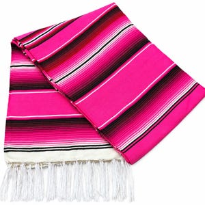 May include: A folded woven blanket featuring a vibrant hot pink base color with horizontal stripes in shades of dark red, black, and white. The textile has a textured appearance and is finished with long, white fringe tassels along the bottom edge.