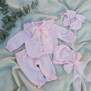May include: A pink knitted baby outfit set with a cardigan, pants, hat, and booties. The cardigan has a lace collar and a bow. The hat also has a lace trim and a bow.
