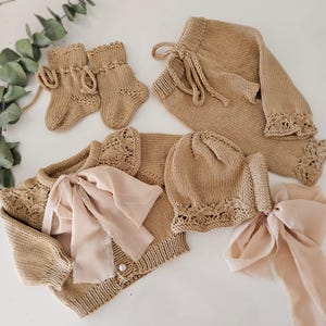 May include: A knitted baby outfit set in a light brown color. The set includes a cardigan with a lace trim, a matching hat, pants, and booties. The cardigan has a bow made of a sheer fabric.