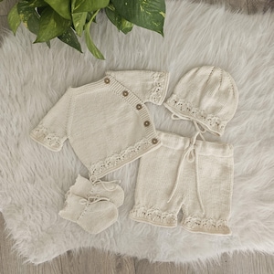 May include: A cream-colored, knitted baby clothing set. The set includes a short-sleeved cardigan with wooden buttons, shorts with a drawstring waist, a bonnet, and booties. All pieces have a decorative lace trim.