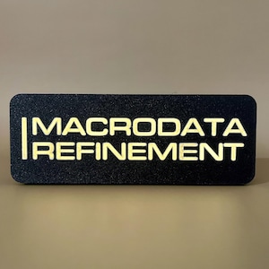 May include: A rectangular black sign with rounded corners. The sign features the words "MACRODATA REFINEMENT" in a bright, glowing yellow font. The text is centered on the sign, which is set against a neutral background.