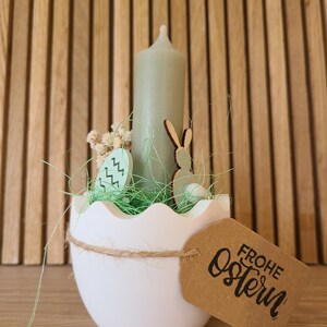 Könnte beinhalten: Ein weißer, eiförmiger Kerzenhalter aus Keramik mit einer grünen Kerze, grünem Gras und einem Holzhasen. Der Halter hat ein braunes Papieretikett mit der Aufschrift "Frohe Ostern" und "Artikel-Nr. 026".