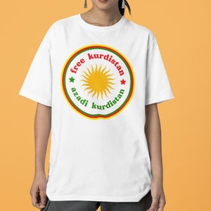 May include: White t-shirt with a circular graphic featuring a yellow sun with red and green stars and the text "free kurdistan azadi kurdistan".