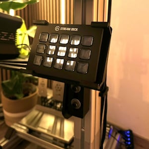 Könnte beinhalten: Ein schwarzes Stream Deck mit 15 Tasten, jede mit einem weißen Licht. Das Stream Deck ist an einer schwarzen Halterung befestigt, die an einer silbernen Stange befestigt ist.