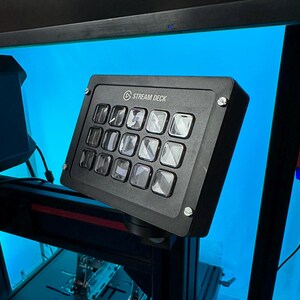 Könnte beinhalten: Ein schwarzes Stream Deck mit 15 Tasten, jede mit einer durchsichtigen Plastikabdeckung. Das Stream Deck ist auf einem schwarzen Ständer montiert.